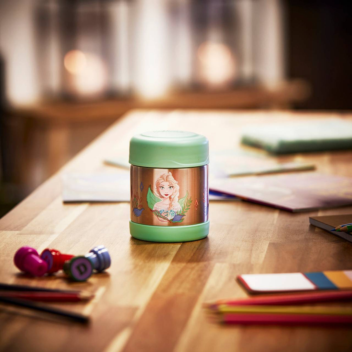 Auf einem Holztisch steht ein grüner Thermobehälter mit einem bunten Bild von einer weiblichen Zeichentrickfigur darauf. Der Hintergrund ist unscharf, und um den Thermobehälter herum liegen verschiedene bunte Schreibutensilien und Bücher.