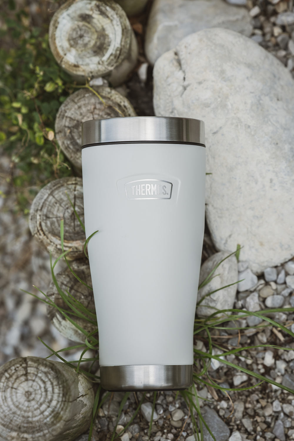 Ein heller THERMOS Isolierbecher liegt zwischen Steinen, Holz und Gras.