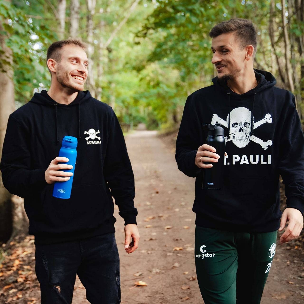 Zwei Männer in schwarzen St.-Pauli-Hoodies laufen durch den Wald und halten THERMOS Flaschen in Blau und Schwarz.