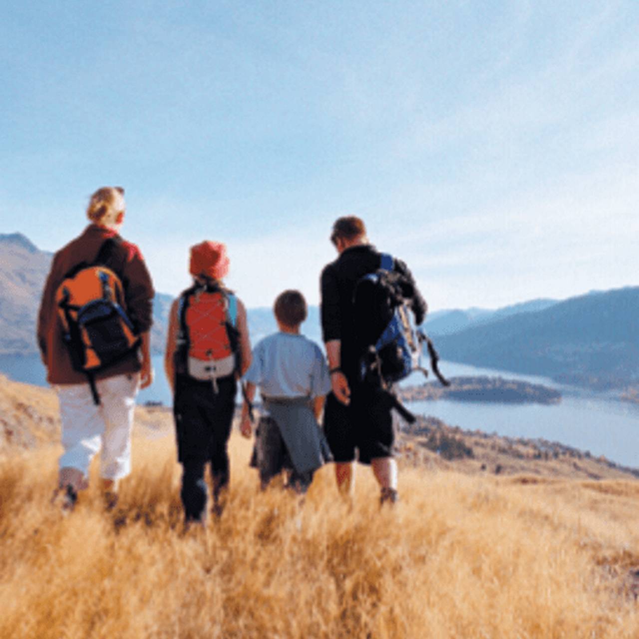 Eine Familie mit Rucksäcken wandert über eine goldene Wiese mit Blick auf einen See und Berge.