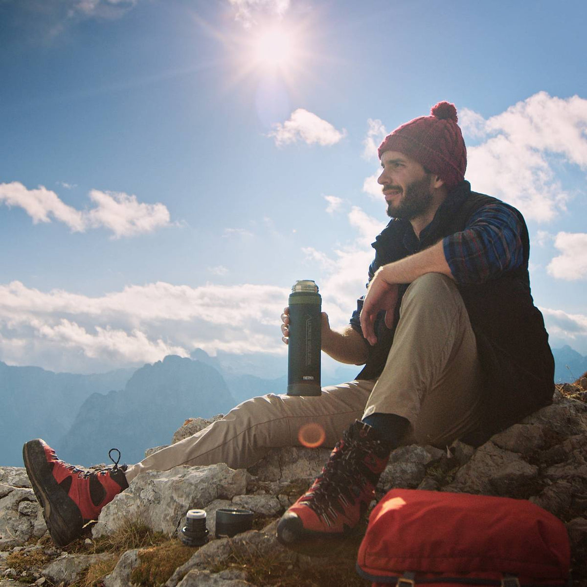 Ein Mann sitzt entspannt auf einem felsigen Berggipfel und hält eine Thermoskanne. Er trägt Wanderausrüstung mit roten Stiefeln und Mütze. Im Hintergrund sieht man Berge und Wolken unter einem blauen Himmel.