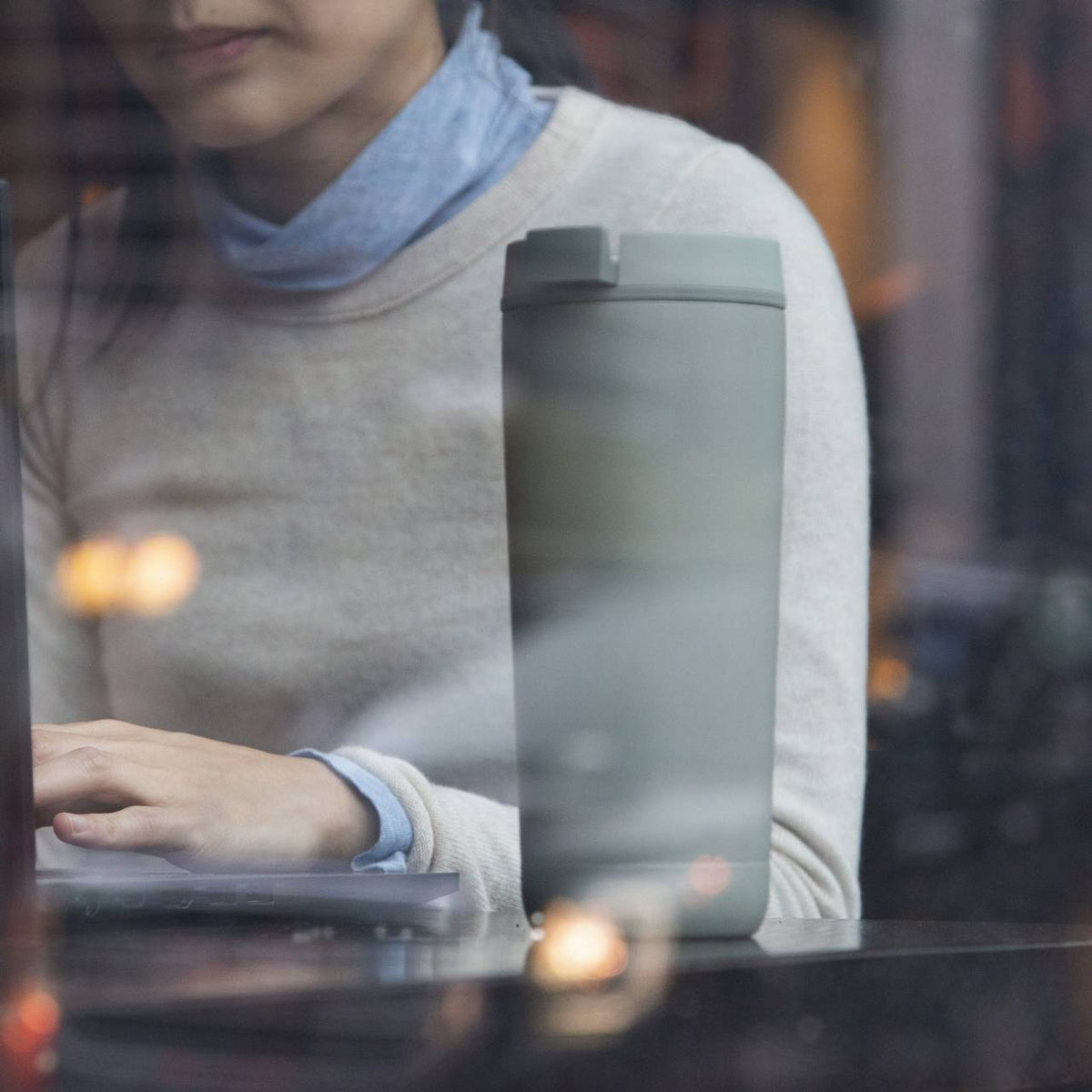 Eine Person sitzt drinnen an einem Laptop, teilweise verdeckt vom Fensterreflexionen. Auf dem Tisch steht ein großer, grauer Thermobecher. Die Person trägt einen hellen Pullover und ein blaues Hemd darunter.