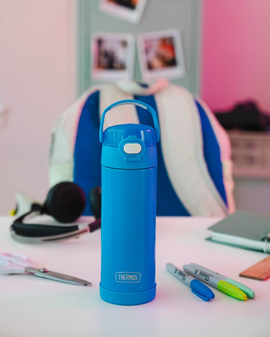 Eine blaue FUNTAINER Kinderflasche steht auf einem Schreibtisch mit Stiften, Kopfhörern und Schulmaterial.