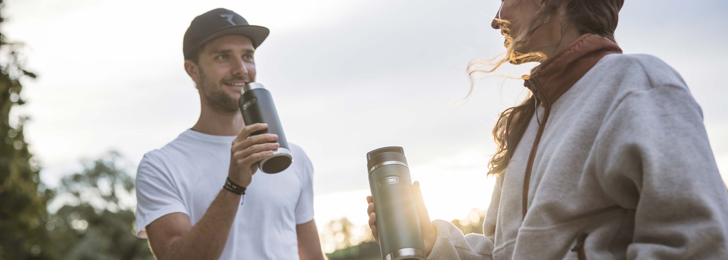 Ein Mann und eine Frau stehen draußen bei Gegenlicht und halten THERMOS Becher in der Hand.