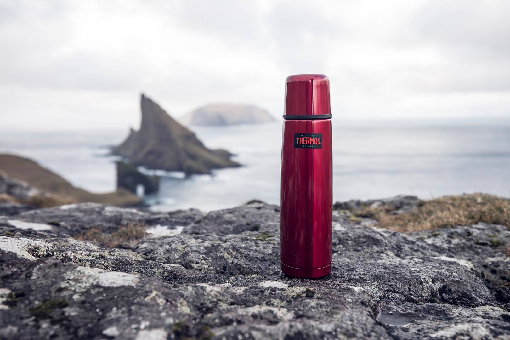 Eine Frau sitzt am Meer im Gras, trinkt aus einem roten Becher und neben ihr steht eine rote Light & Compact Thermosflasche.