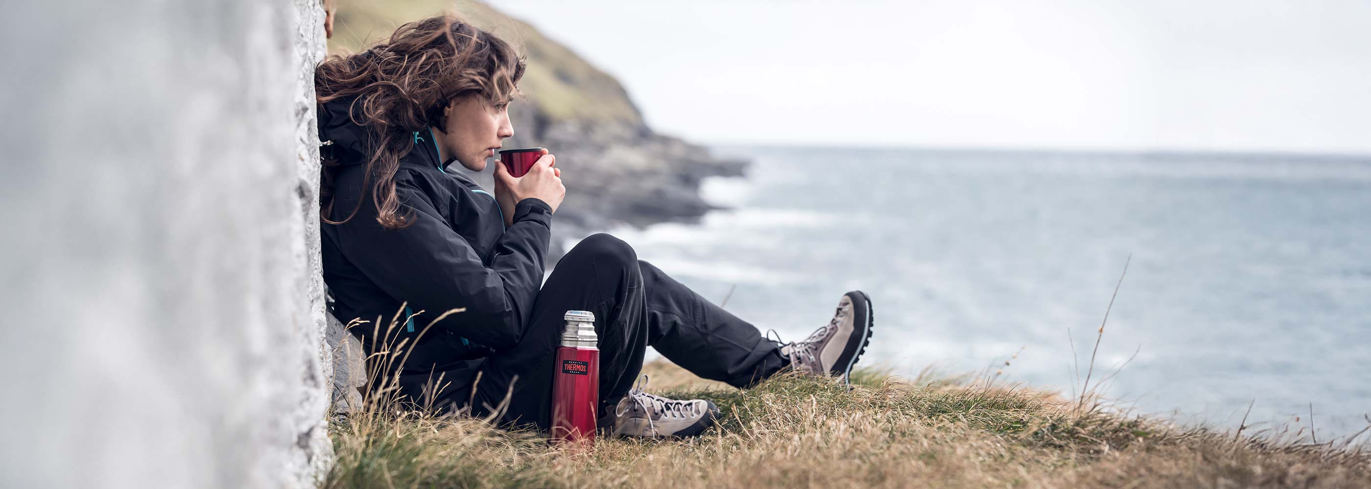 Eine rote Light & Compact Thermosflasche steht auf einem Felsen vor einer Küstenlandschaft mit Meer und Felsen.