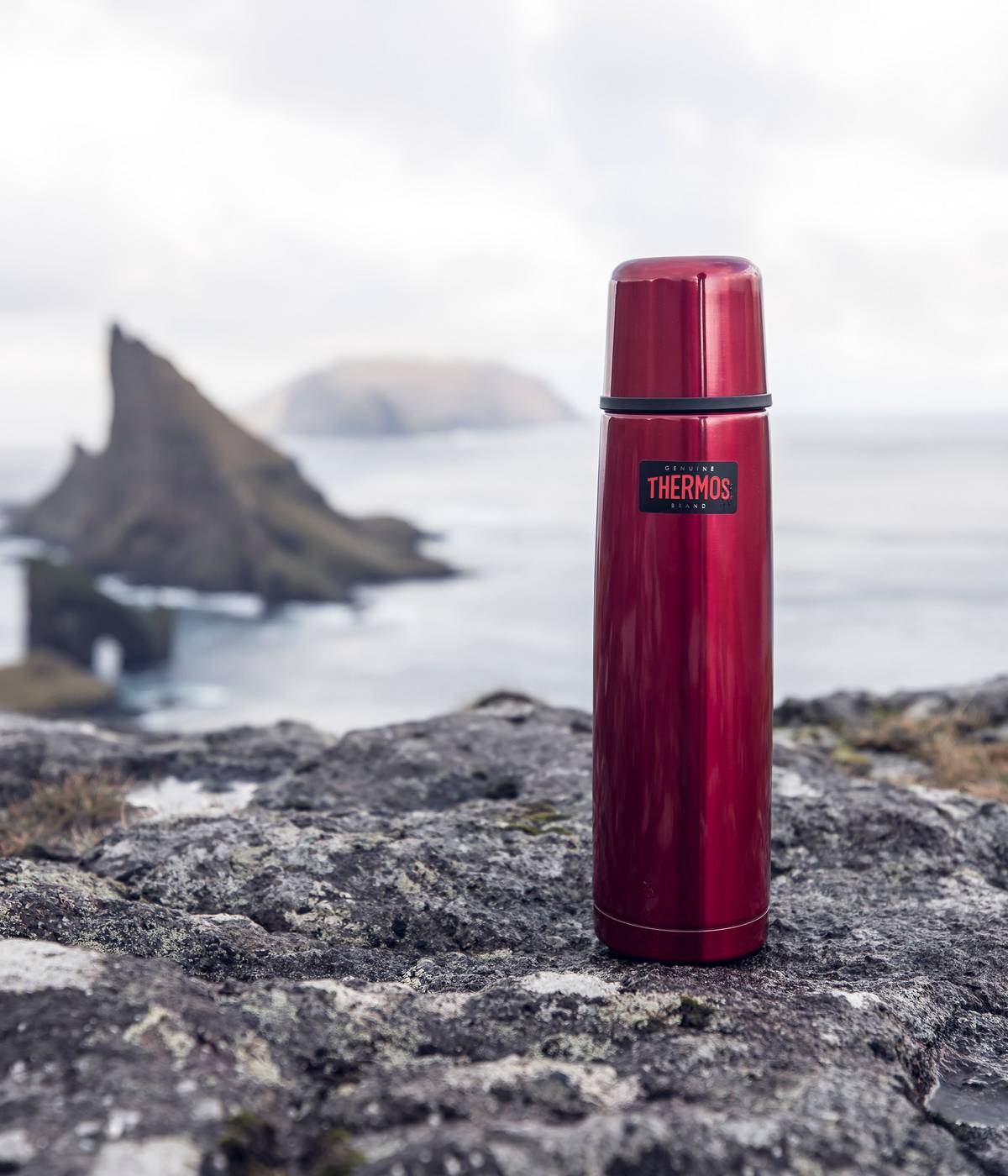 Eine rote Light & Compact Thermosflasche steht auf einem Felsen vor einer Küstenlandschaft mit Meer und Felsen.