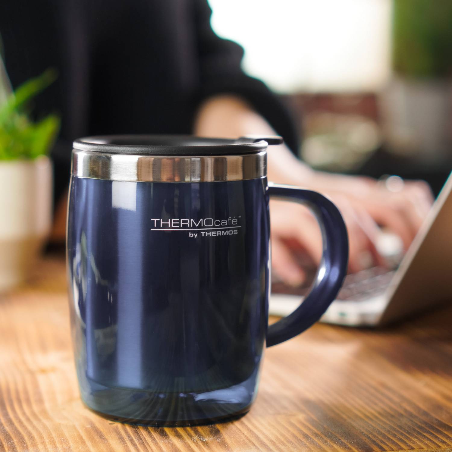 Nahaufnahme eines blauen THERMOcafé Desktop Cups mit Deckel auf einem Holztisch; im Hintergrund tippt eine Person am Laptop.