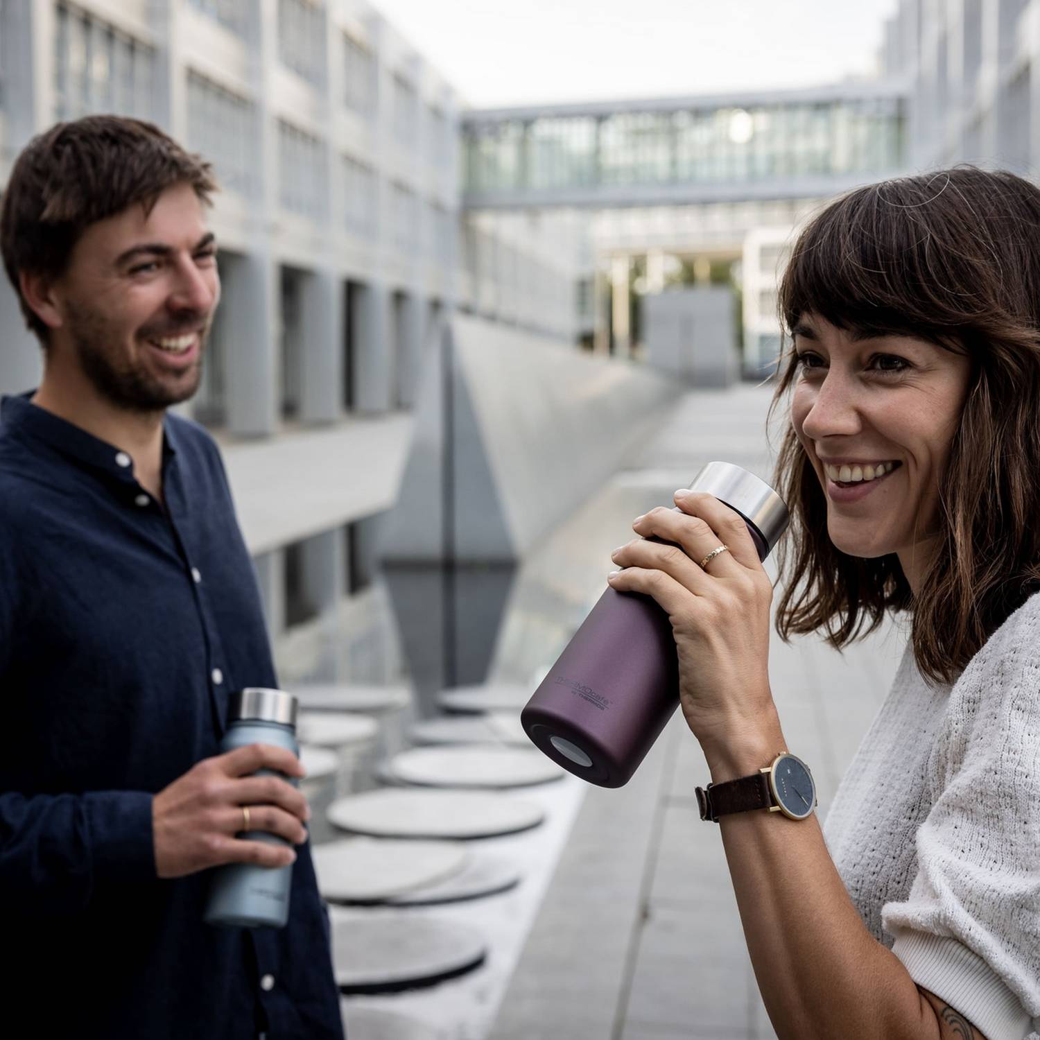 Eine Frau trinkt aus einer lilafarbenen THERMOcafé Flasche, während ein Mann mit einer blauen Flasche neben ihr steht.