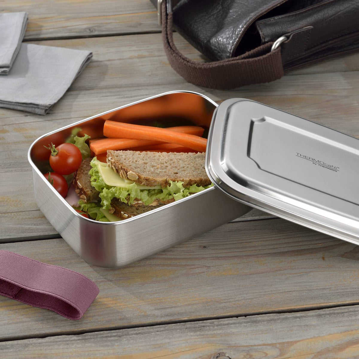 Eine Lunchbox mit Sandwich, Radieschen und Gurkenscheiben steht auf einem Tisch; im Hintergrund schreibt eine Person in ein Notizbuch.