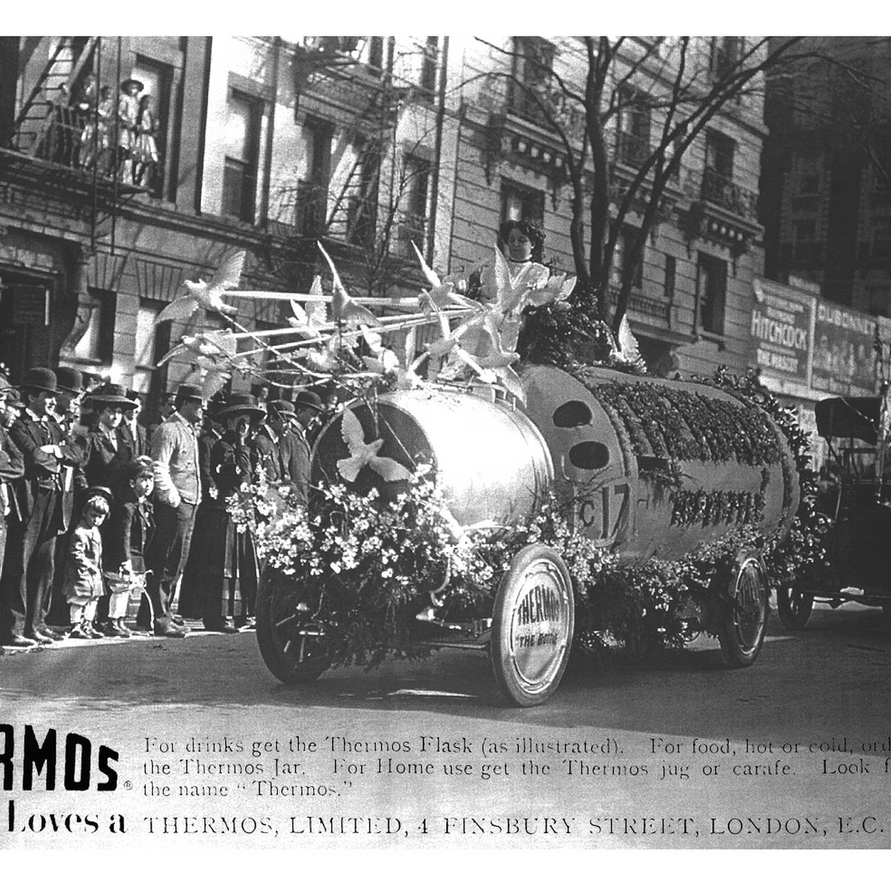Historische Schwarz-Weiß-Aufnahme eines dekorierten Werbewagens in Form einer Thermosflasche bei einer Parade mit Zuschauern.